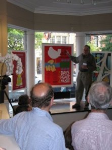 Man stands on stage in front of crowd, gesturing at poster; blue, beige, red, grey